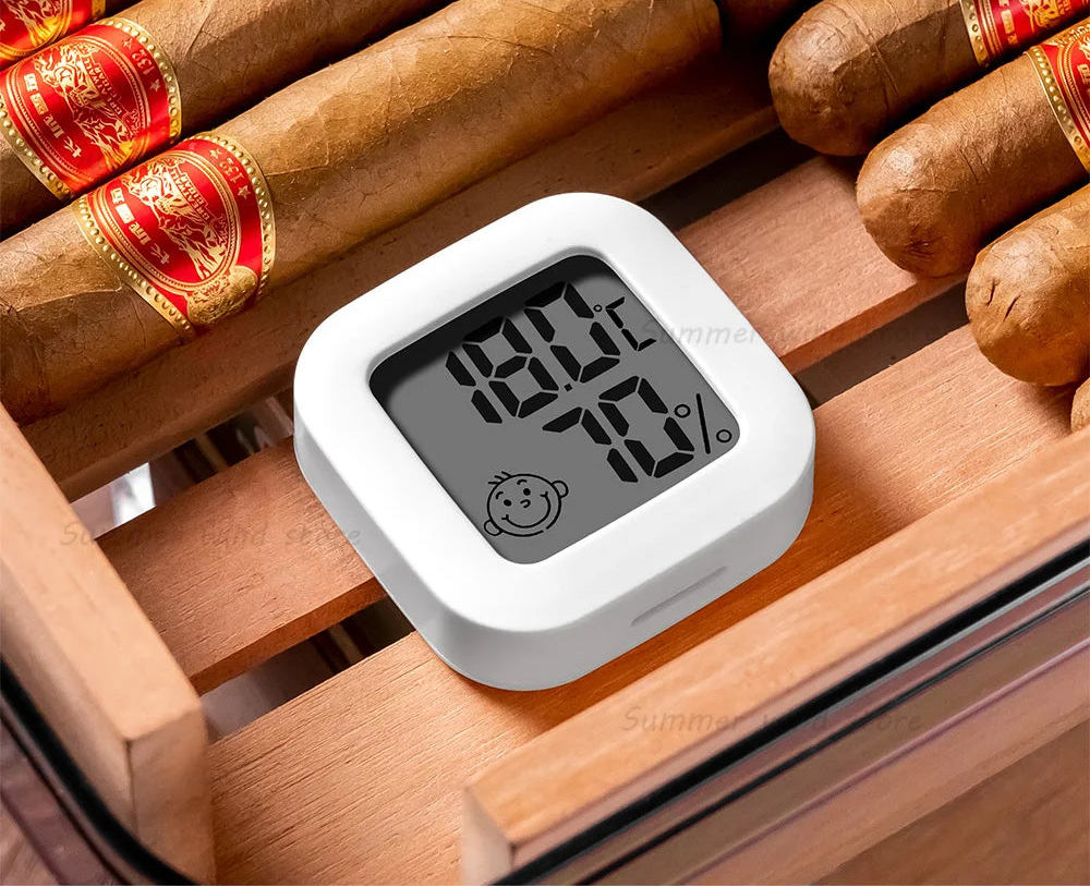 Digital hygrometer on a wooden surface with cigars, displaying temperature and humidity levels.