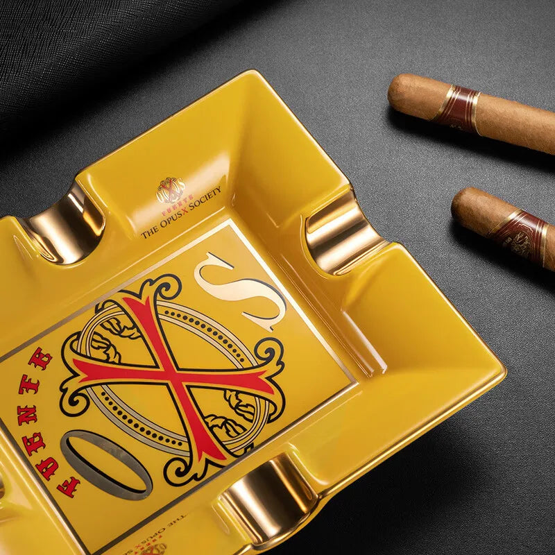 Yellow cigar ashtray with a opusx logo and two cigars on a dark surface